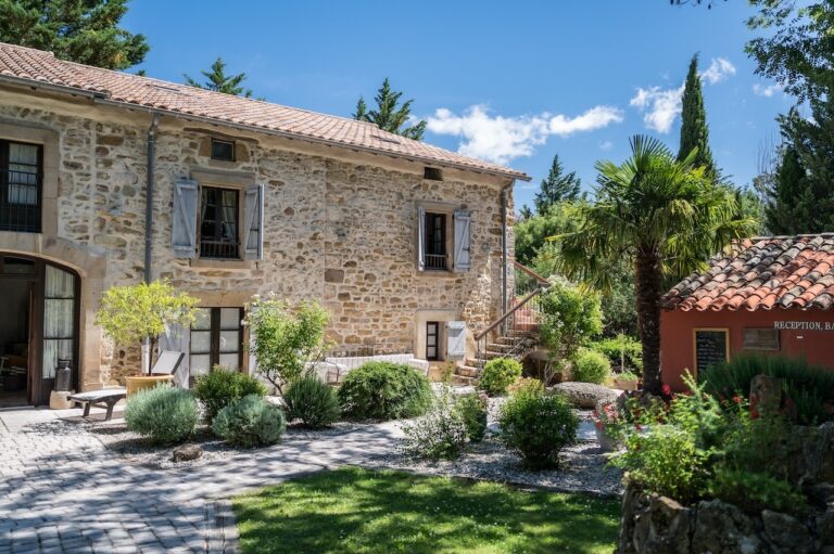 View of traditional French stone farmhouse in the background with gravel driveway and shrubs in the foreground